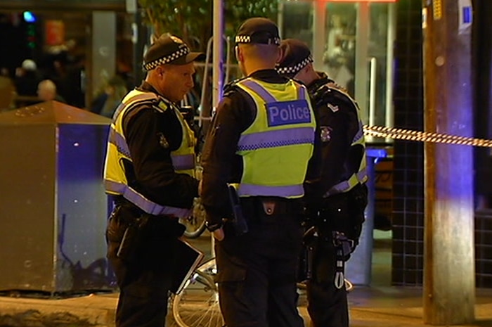 Three police officers speak to each other in front of a scene marked by police tape.