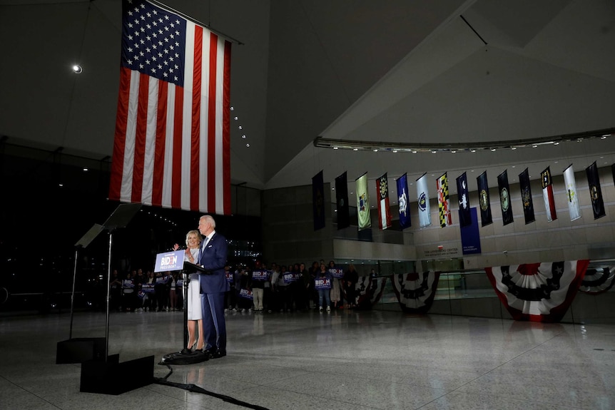 Joe Biden and his wife Jill stand at a podium in a large room with a long American flag hanging above them.