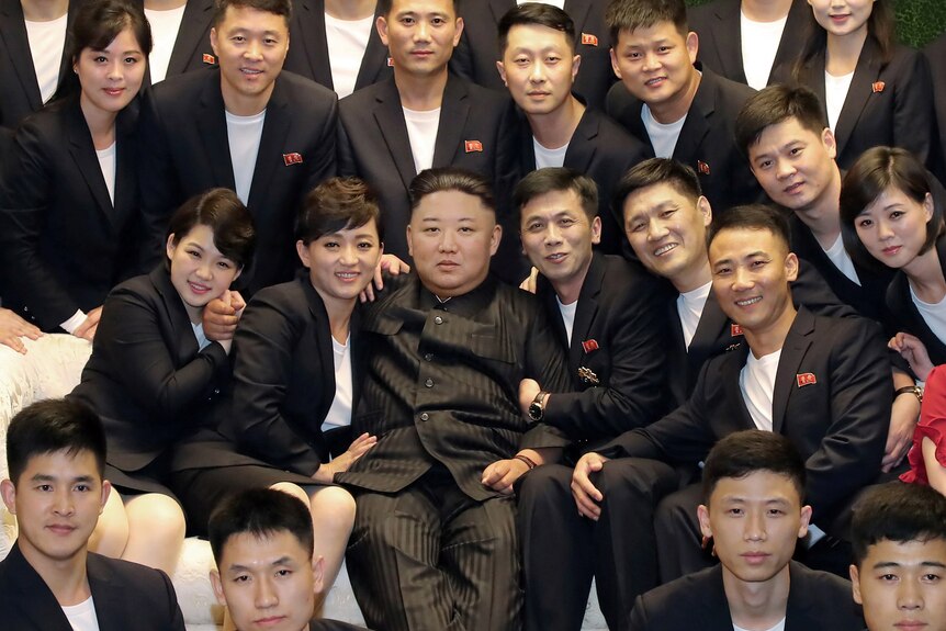 Kim Jong Un on a couch surrounded by people in dark suits and white t-shirts