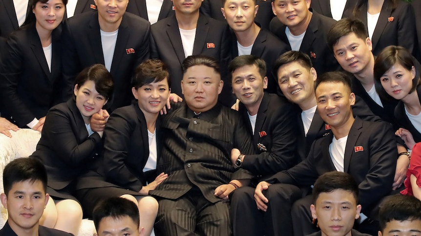Kim Jong Un on a couch surrounded by people in dark suits and white t-shirts