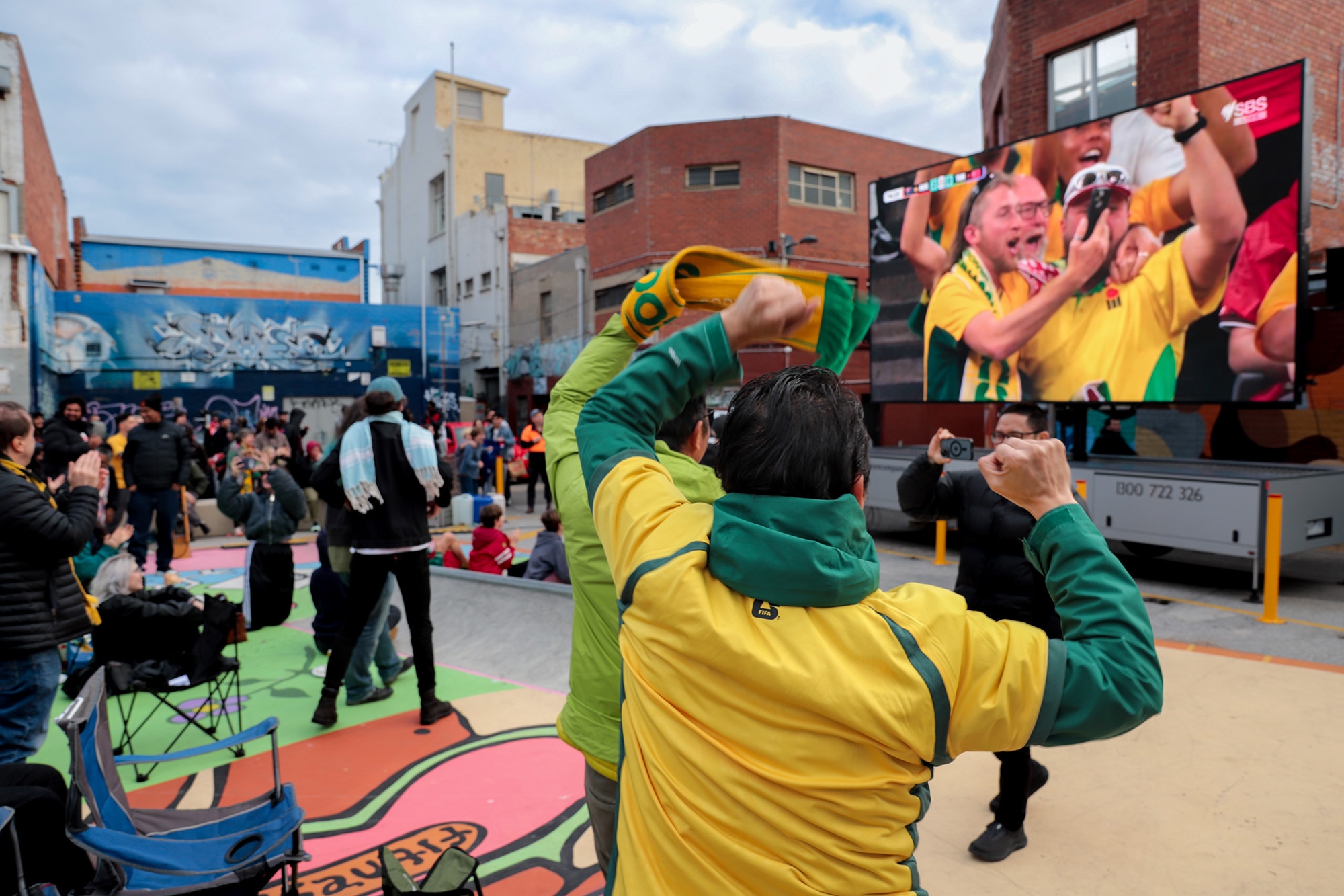 Seen from behind, men wearing green and gold shirts and one holding a scarf, raise their arms as they look at a big screen in f 