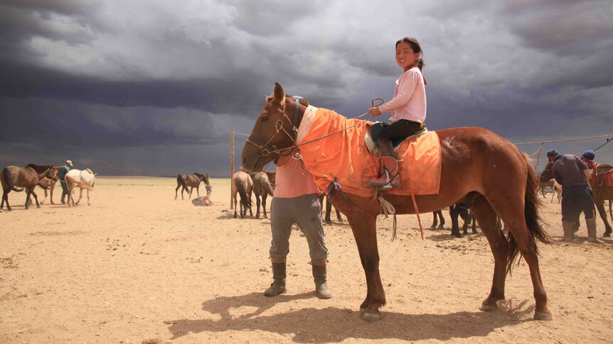 A young girl sits on a horse in Mongolia as dark storm clouds sit in the background.