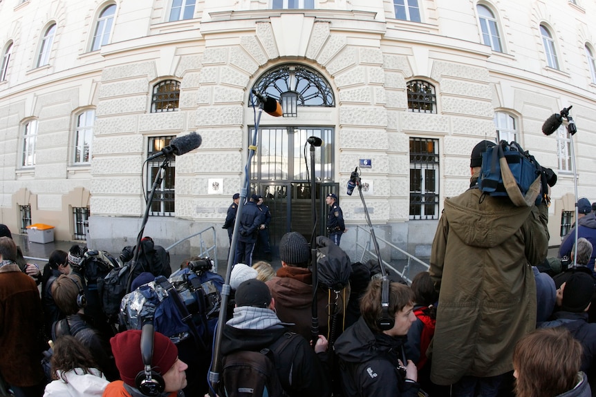 A group of cameras and reporters stand outside a white brick building 