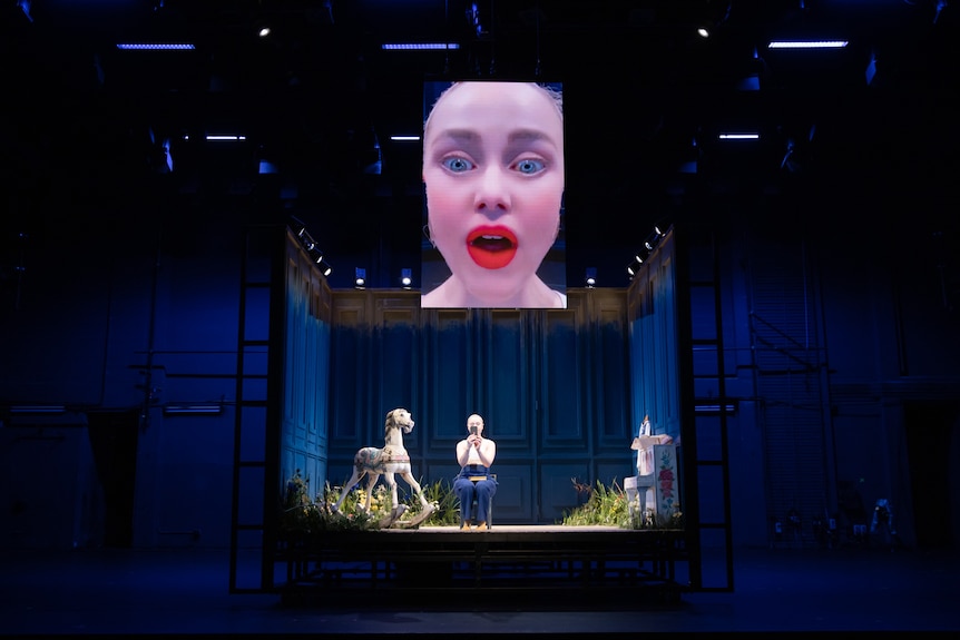 White actress wears wig cap, white shirt and blue pants and sits on stage holding phone while above a screen projects her face.