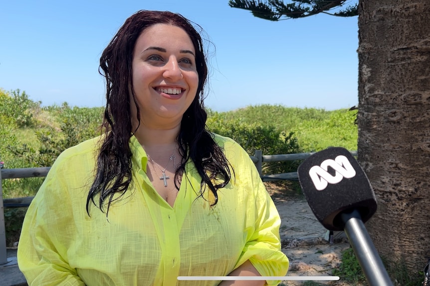 Woman with dark hair, wearing yellow shirt, smiling in front of blue sky and ABC microphone