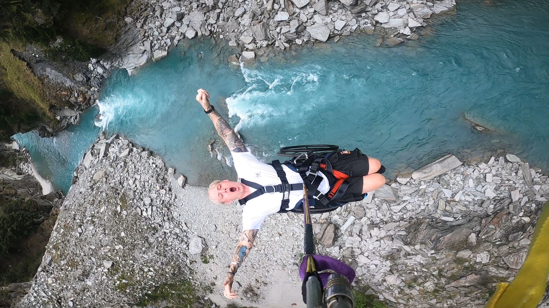 A man in a wheelchair hanging from a canyon swing rope above a crystal blue river