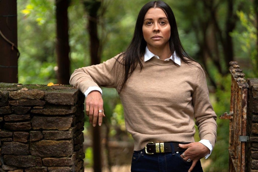 An unsmiling woman with long black hair leanes on a stone fence in a gateway.