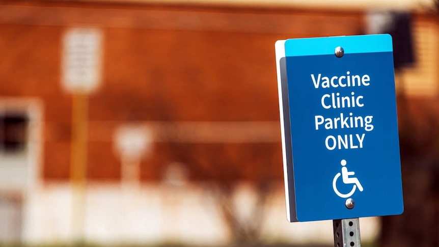 Blue sign reads "vaccine clinic disabled parking only" against the blurred backdrop of a carpark.