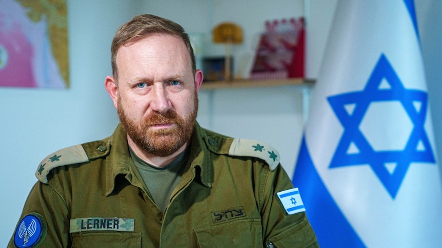 A man in an army uniform inside an office looks at the camera with a neutral expression. In the background, a flag is visible