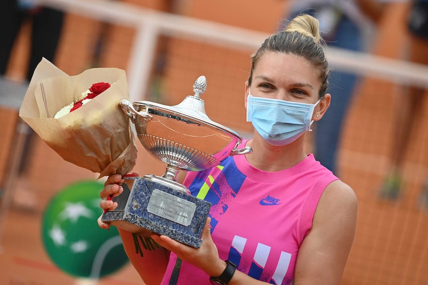 Simona Halep holds a trophy and a bunch of flowers while wearing a mask