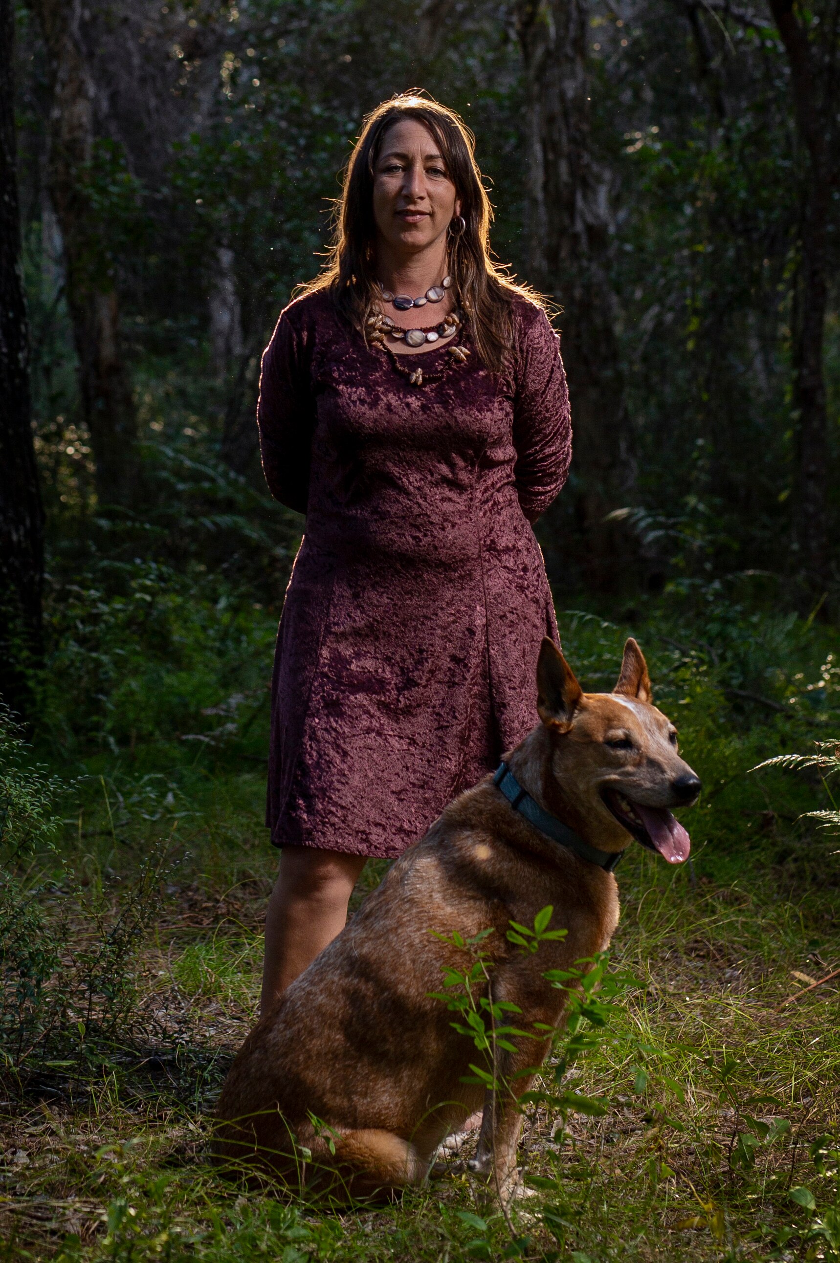 A long-haired brunette Aboriginal woman stands with her arms behind her back in dense forest, a dog sitting at her feet