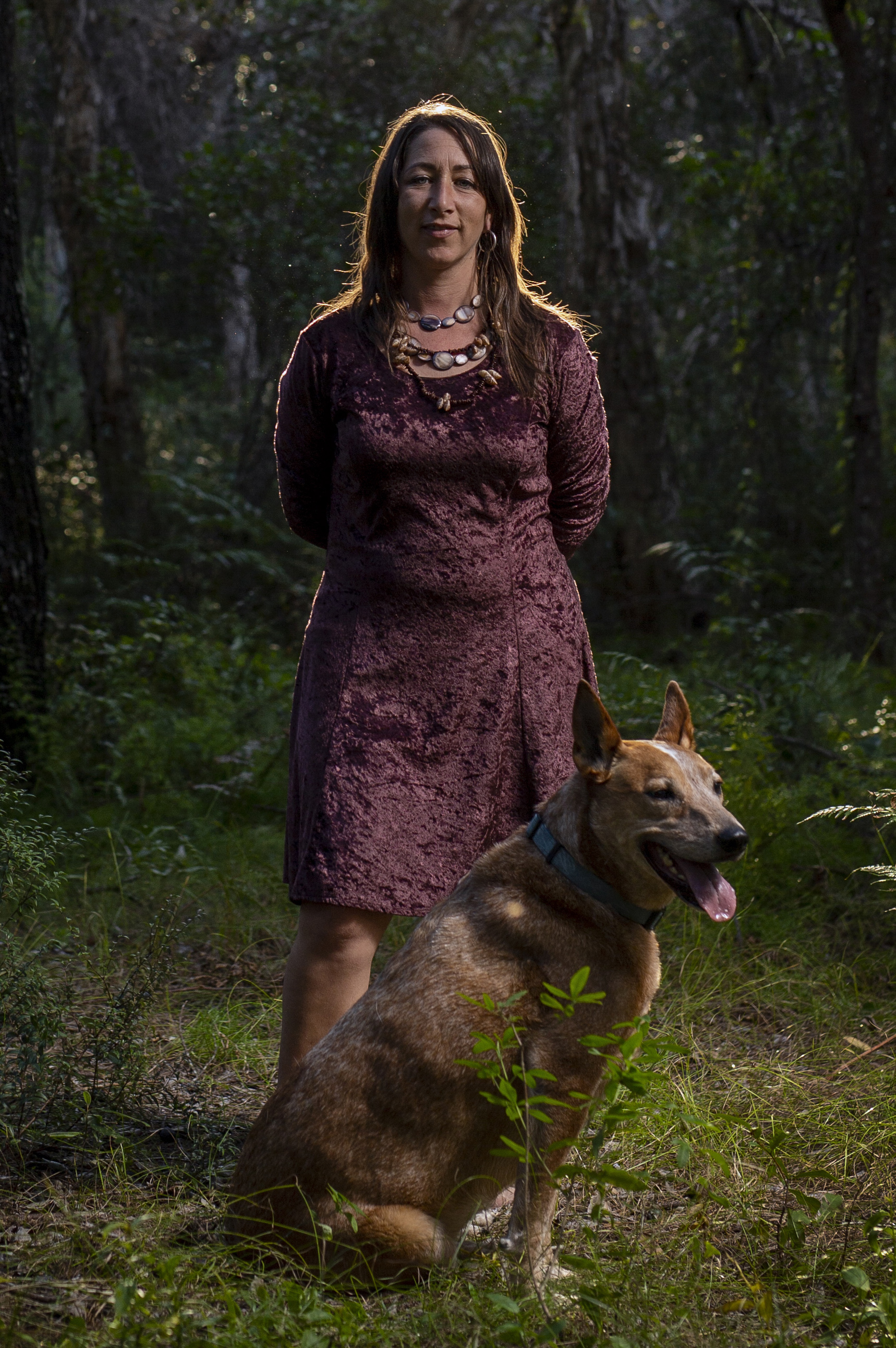 A long-haired brunette Aboriginal woman stands with her arms behind her back in dense forest, a dog sitting at her feet