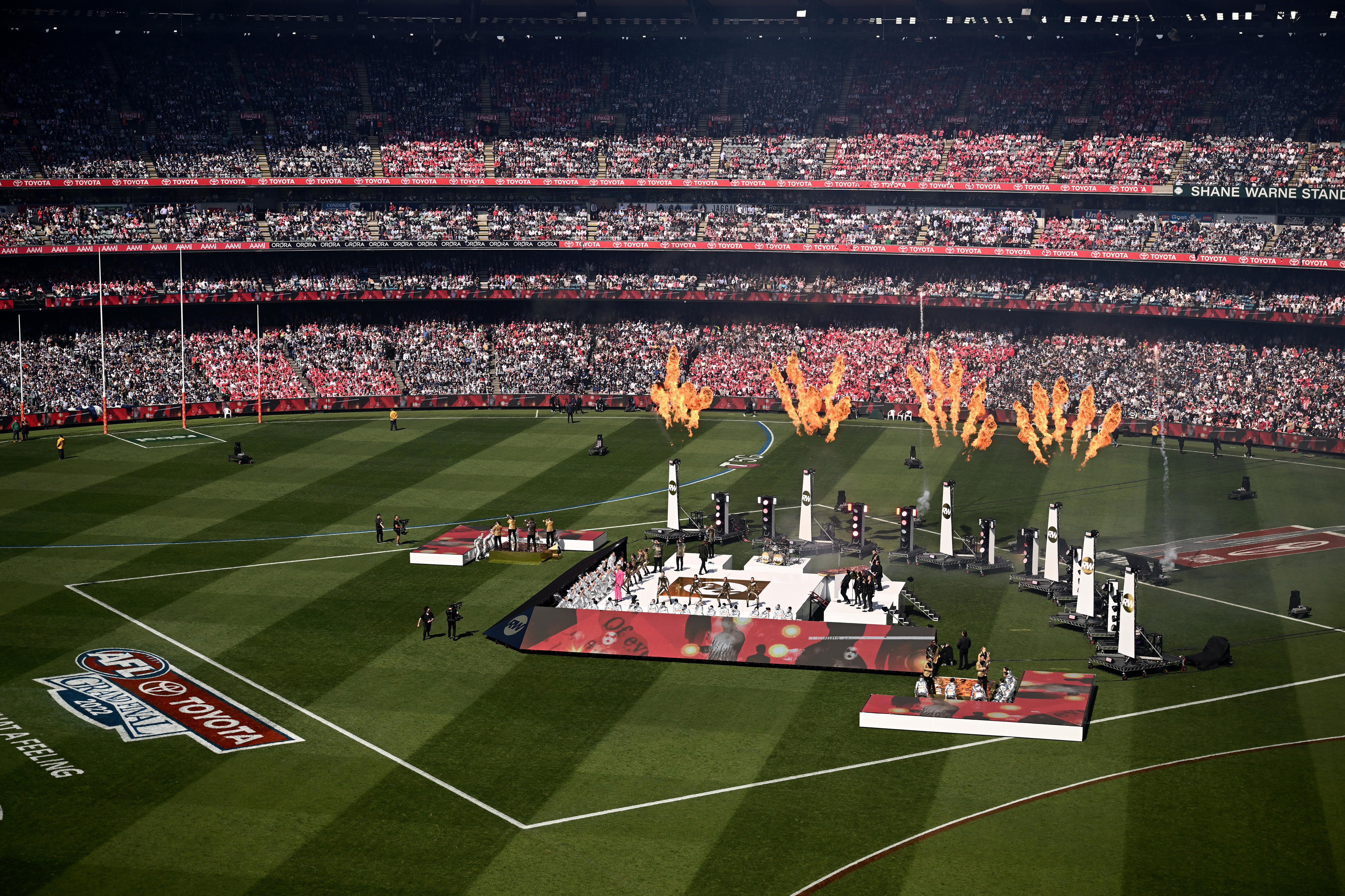Wide shot of Robbie Williams performing on stage in the middle of the MCG with fireworks behind him.