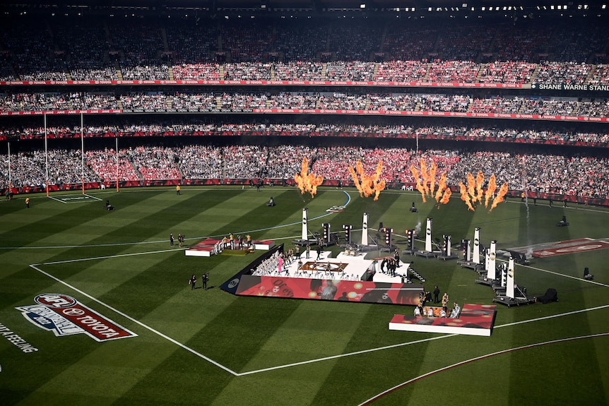 A wide shot of Robbie Williams performing on a stage in the middle of the MCG, with pyrotechnics behind him.