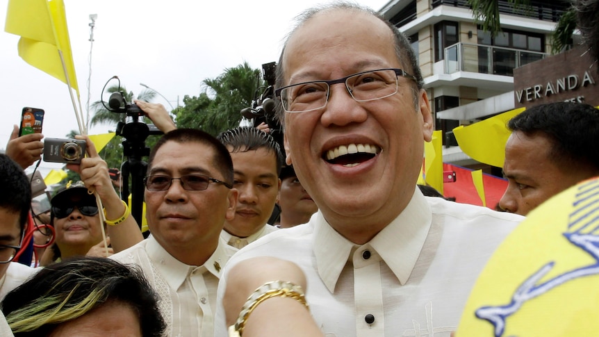 A shot from the shoulders up of Benigno Aquino smiling as he is welcomed by a crowd of supporters. 