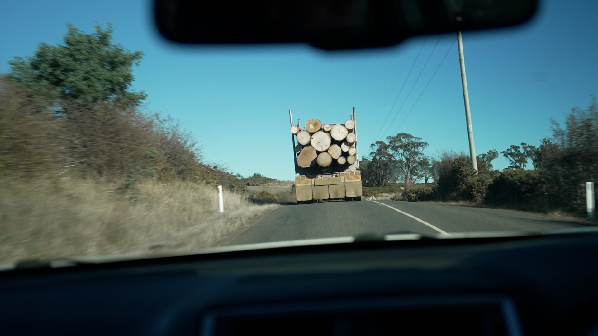 A photo shot from through the windshield of a car of a country road. Ahead is the back of a truck loaded with large logs.