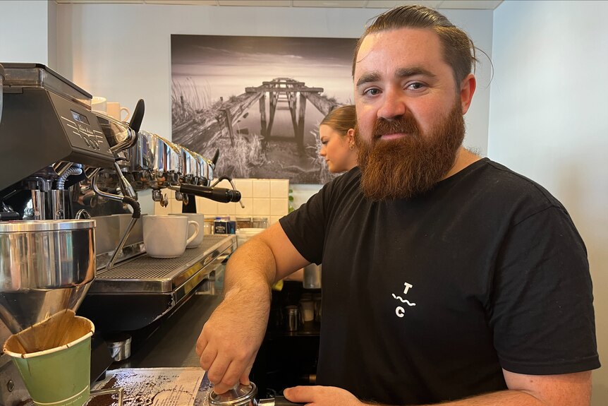 A bearded man stands beside a coffee machine.