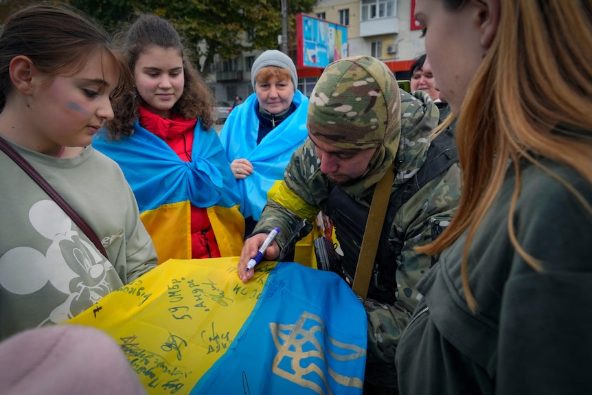 A Ukrainian soldier writes: "From the Ukrainian Armed Forces" on the national flag he signs for a young woman.
