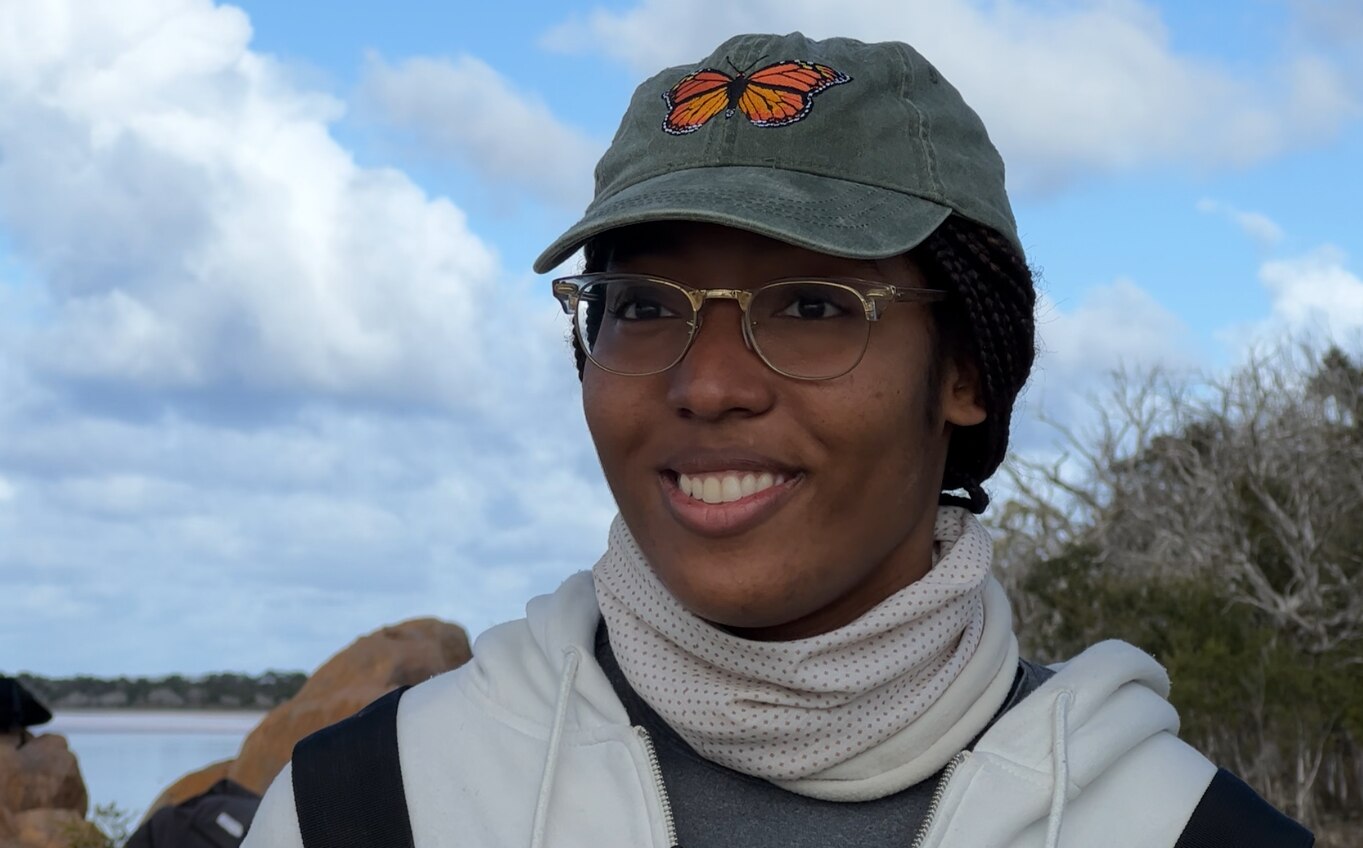 A smiling African-American woman, wears a cap with an embroidered butterfly, a scarf around her neck and glasses. Lake behind.