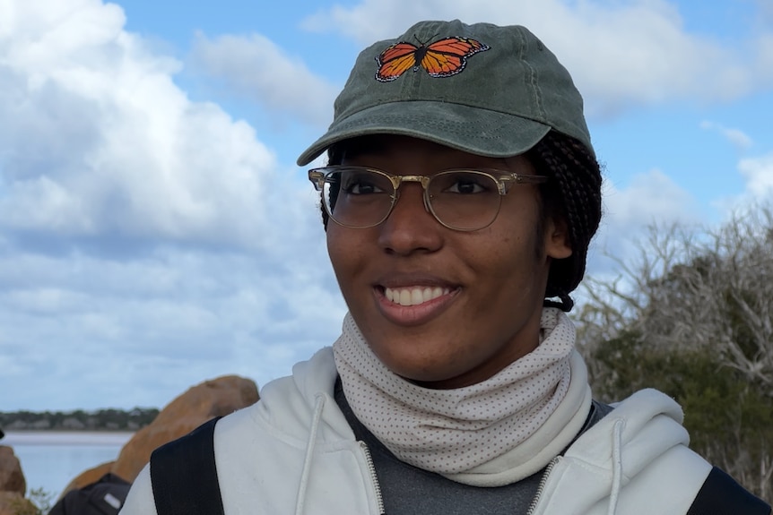 A smiling African-American woman, wears a cap with an embroidered butterfly, a scarf around her neck and glasses. Lake behind.