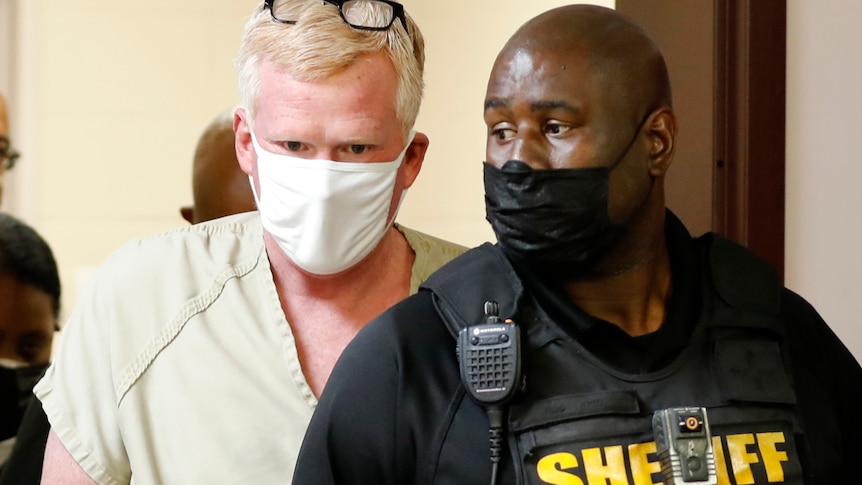 Man in prison outfit walks through a doorway with a sheriff in a vest in front of him 