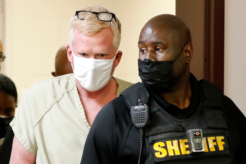 Man in prison outfit walks through a doorway with a sheriff in a vest in front of him 