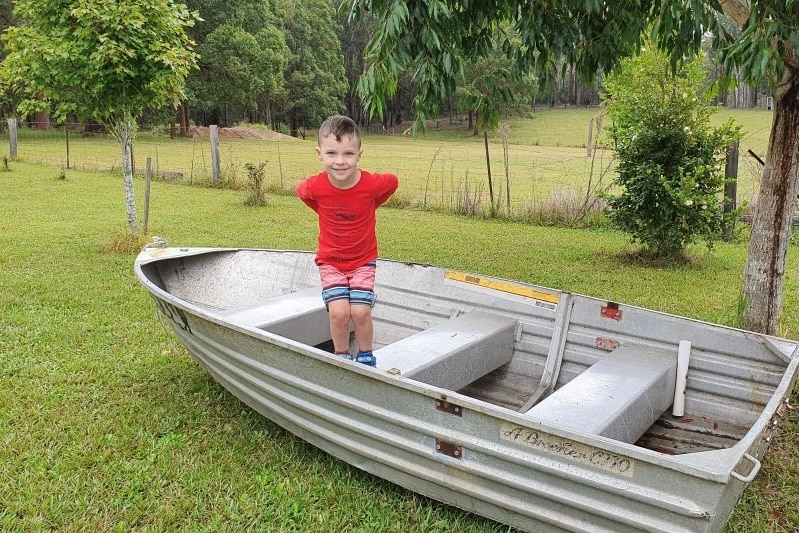 A three-year-old boy, wearing a red T-shirt, stands in a tinny on dry land