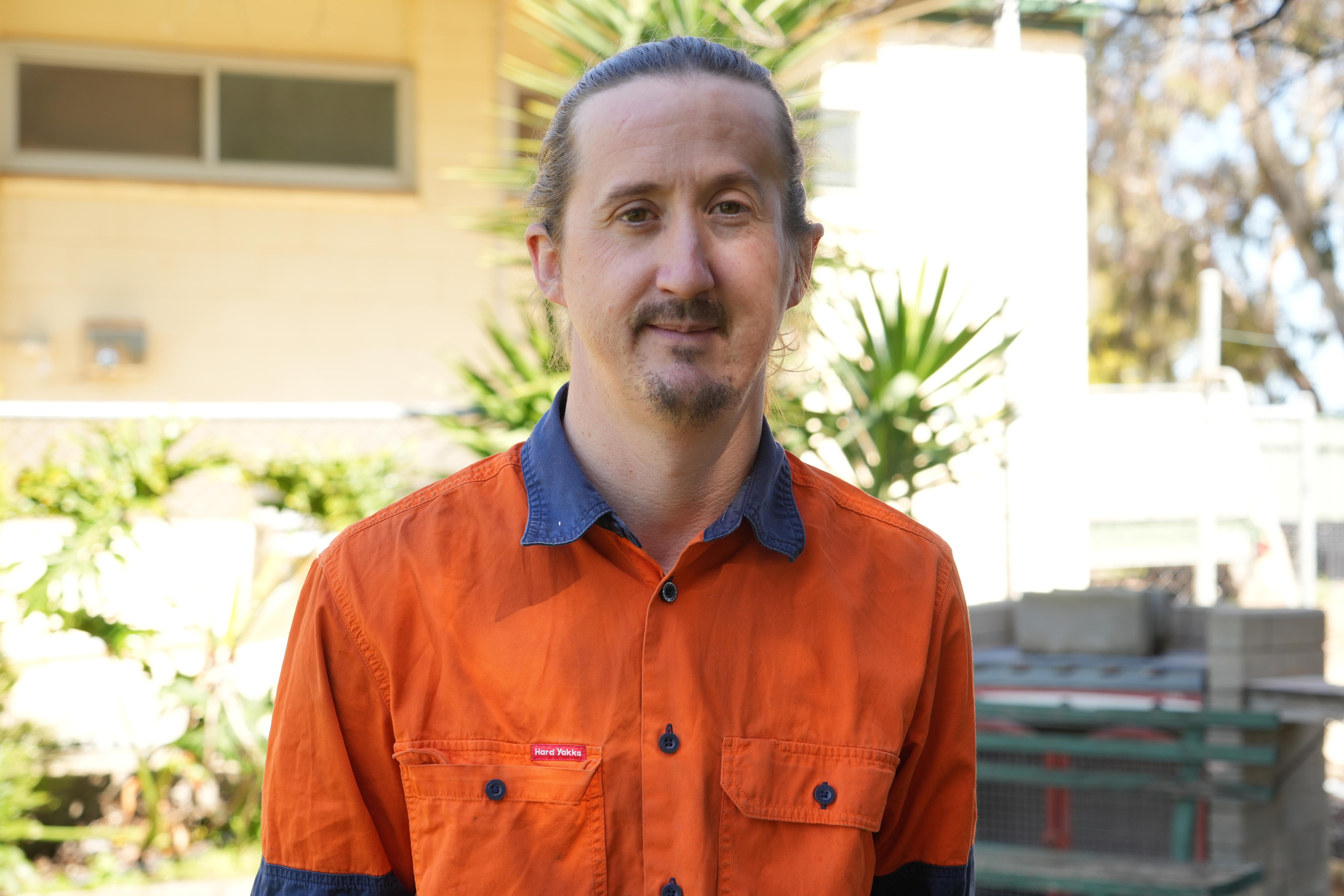A man with a goatie and tied back hair in an orange work shirt, Haiden Button, standing in front of a building and shrubs.