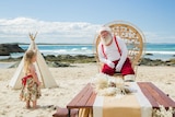 Santa sitting on a chair on the beach, talking to a small girl standing nearby.