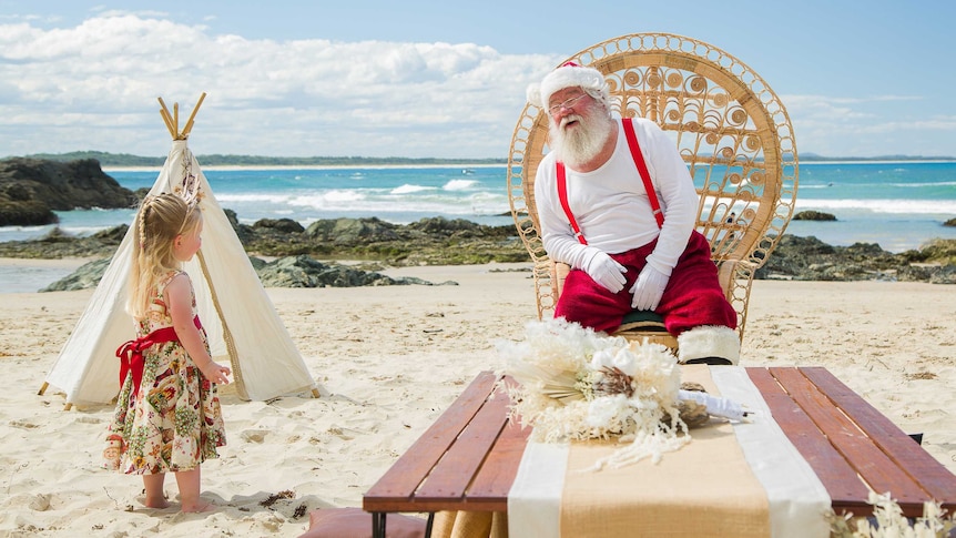 Santa sitting on a chair on the beach, talking to a small girl standing nearby.