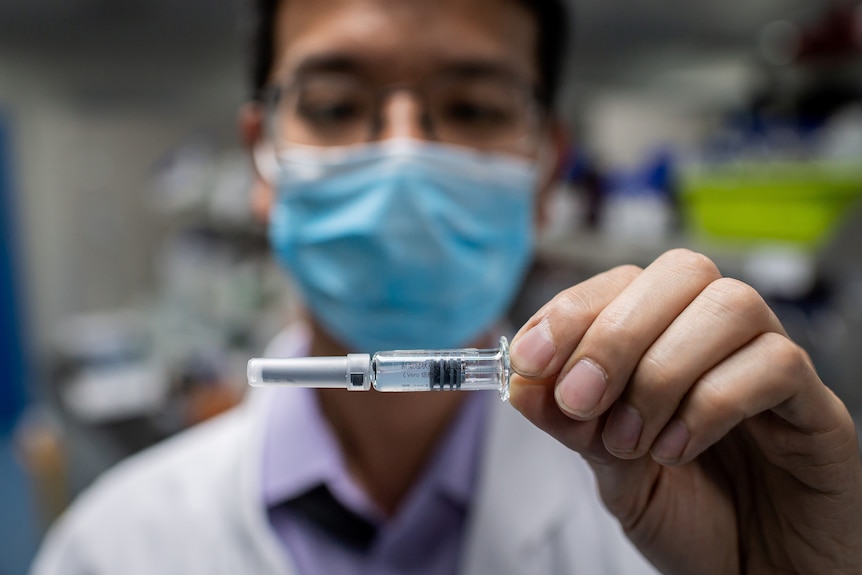 A man wearing a blue face mask holds up a vial of experimental COVID-19 vaccine.