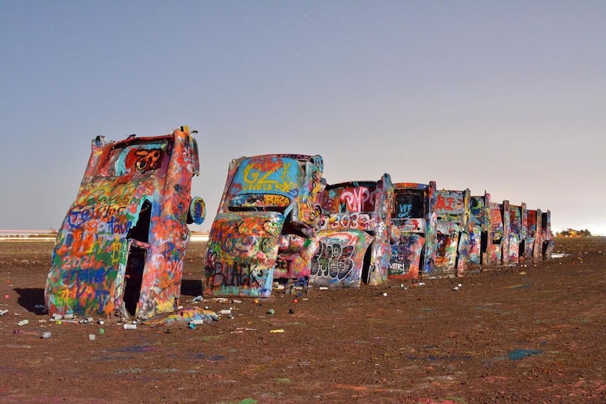 Ten Cadillac cars covered in bright graffiti are half-buried in dirt.