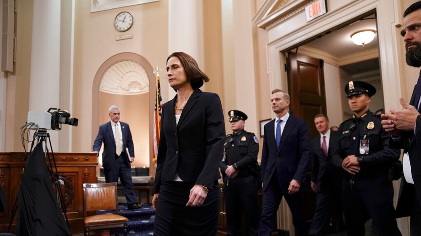 A man and woman in dark suits walk through a US Congress chamber surrounded by police and television cameras.