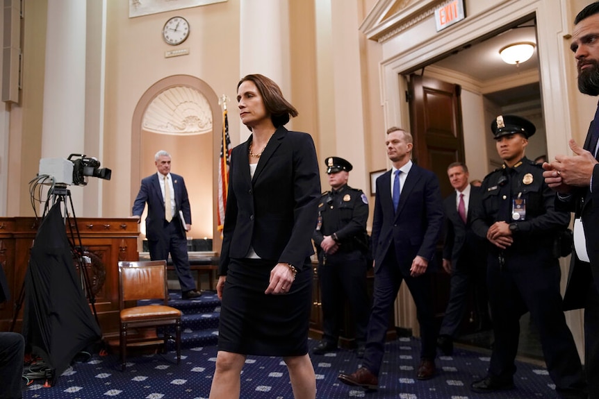 A man and woman in dark suits walk through a US Congress chamber surrounded by police and television cameras.