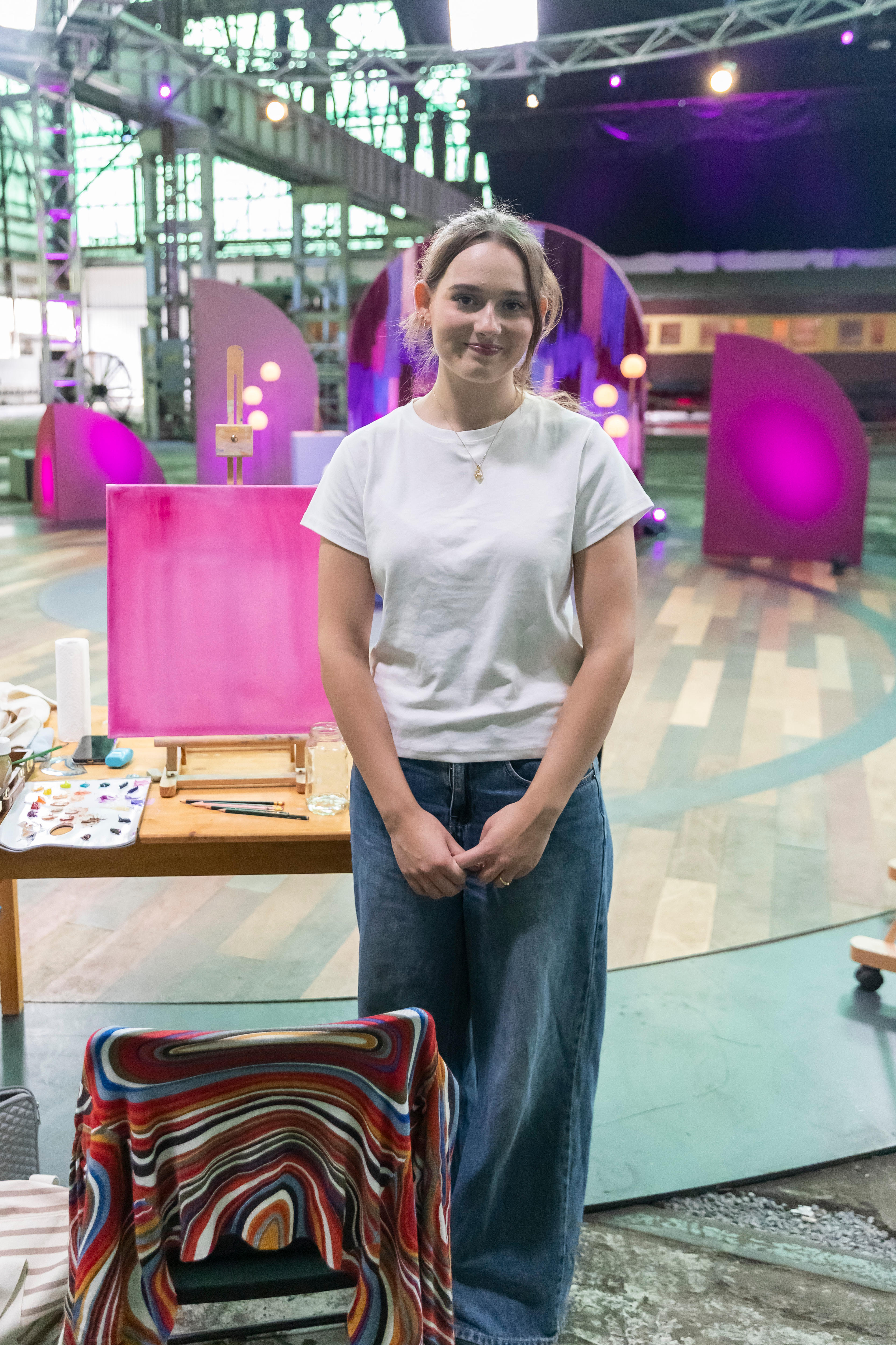 A young woman with brown hair smiles at the camera. She wears a white t-shirt. Behind her are pink shapes on a TV set