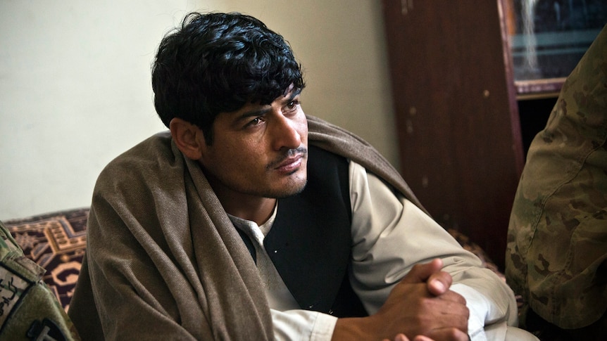 A man wearing Afghan clothing sits indoors listening intently.