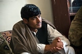 A man wearing Afghan clothing sits indoors listening intently.