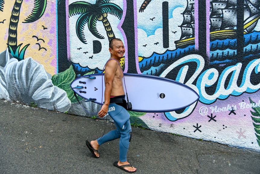 A Japanese man carrying a surfboard and walking down the street.