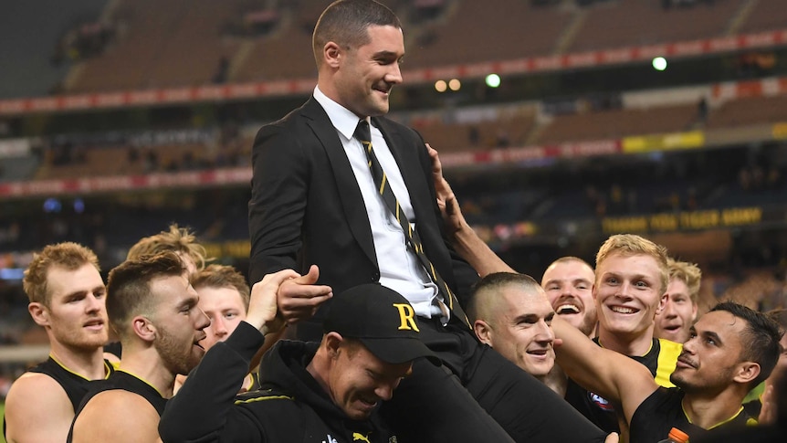 A group of smiling Richmond players carry Shaun Grigg — who is wearing a suit — off the MCG.