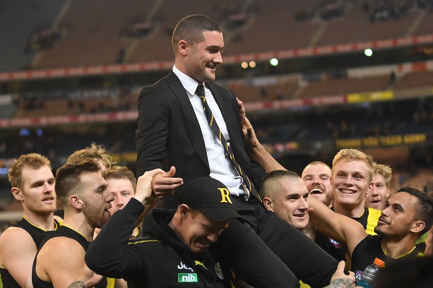 A group of smiling Richmond players carry Shaun Grigg — who is wearing a suit — off the MCG.