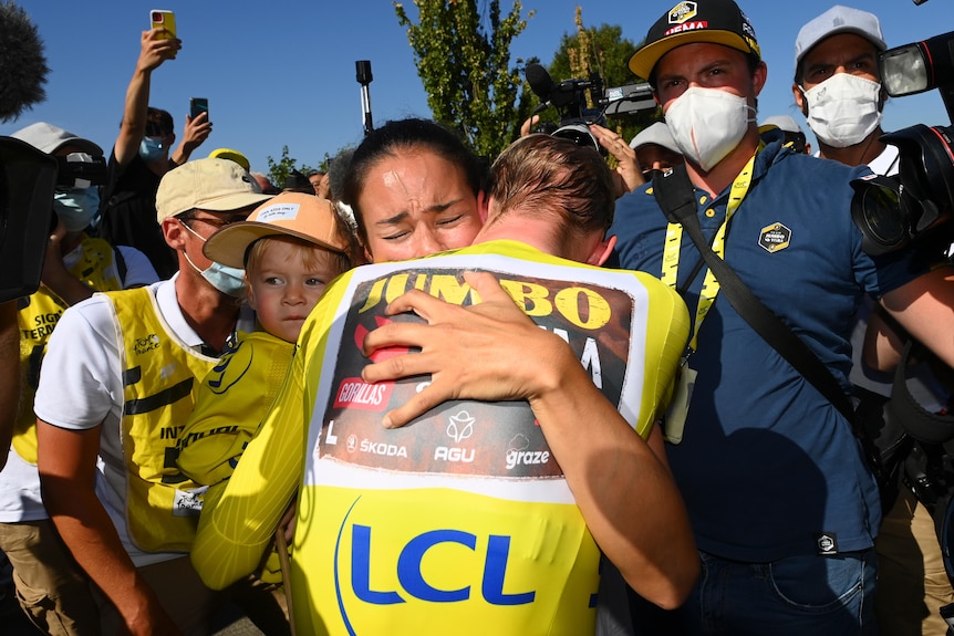 A woman with tears in her eyes hugs her husband in a yellow jersey as he holds their child after a Tour de France stage.