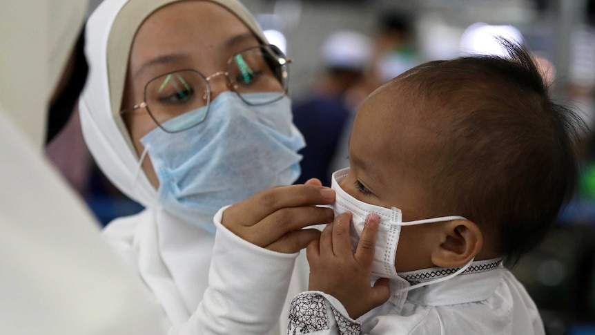 A woman in white with large glasses fits a mask on a child in Malaysia.