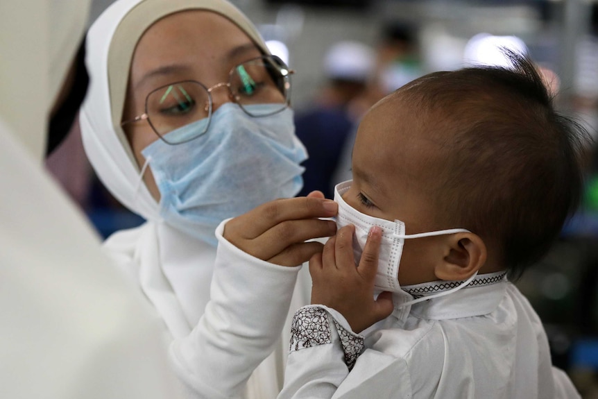 A woman in white with large glasses fits a mask on a child in Malaysia.