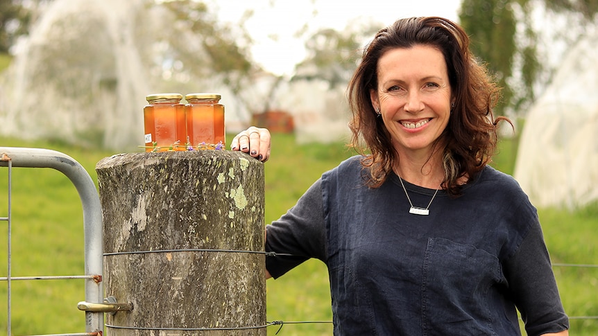 Deb McLaughlin leans on a fence post next to two jars of honey with bee hives and fruit trees in the background.