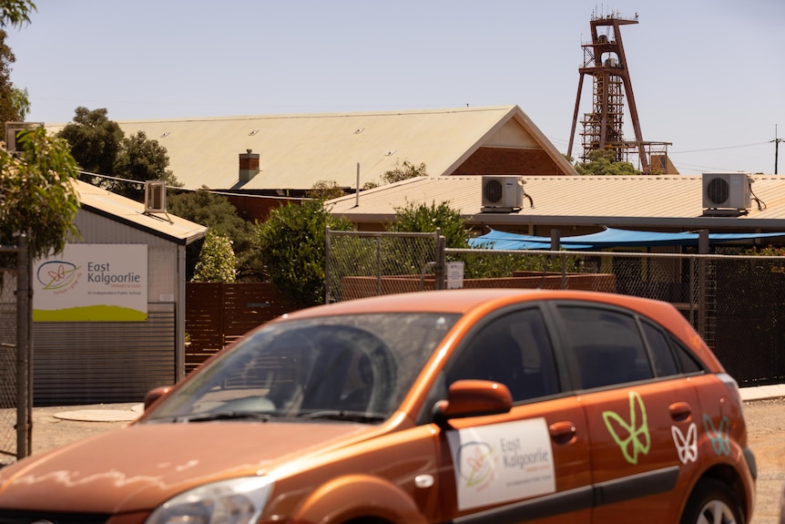 A car parked out the front of a school with a gold mine headframe in the background. 