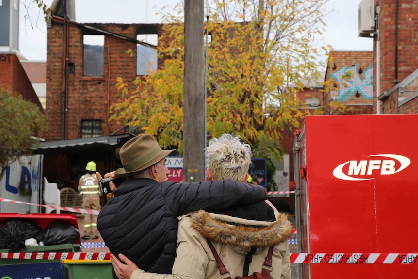 Two people with their backs to the camera embrace in front of La Mama Theatre, which was destroyed by fire.