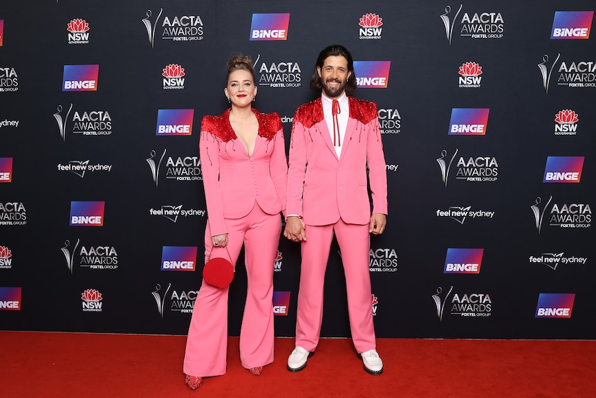 A blonde white woman and brunette white man wear matching hot pink suits with glittery red shoulder pads on a red carpet.