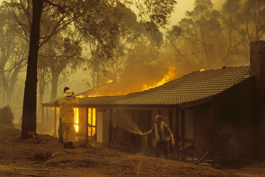 A man holds a hose while a home behind him burns.