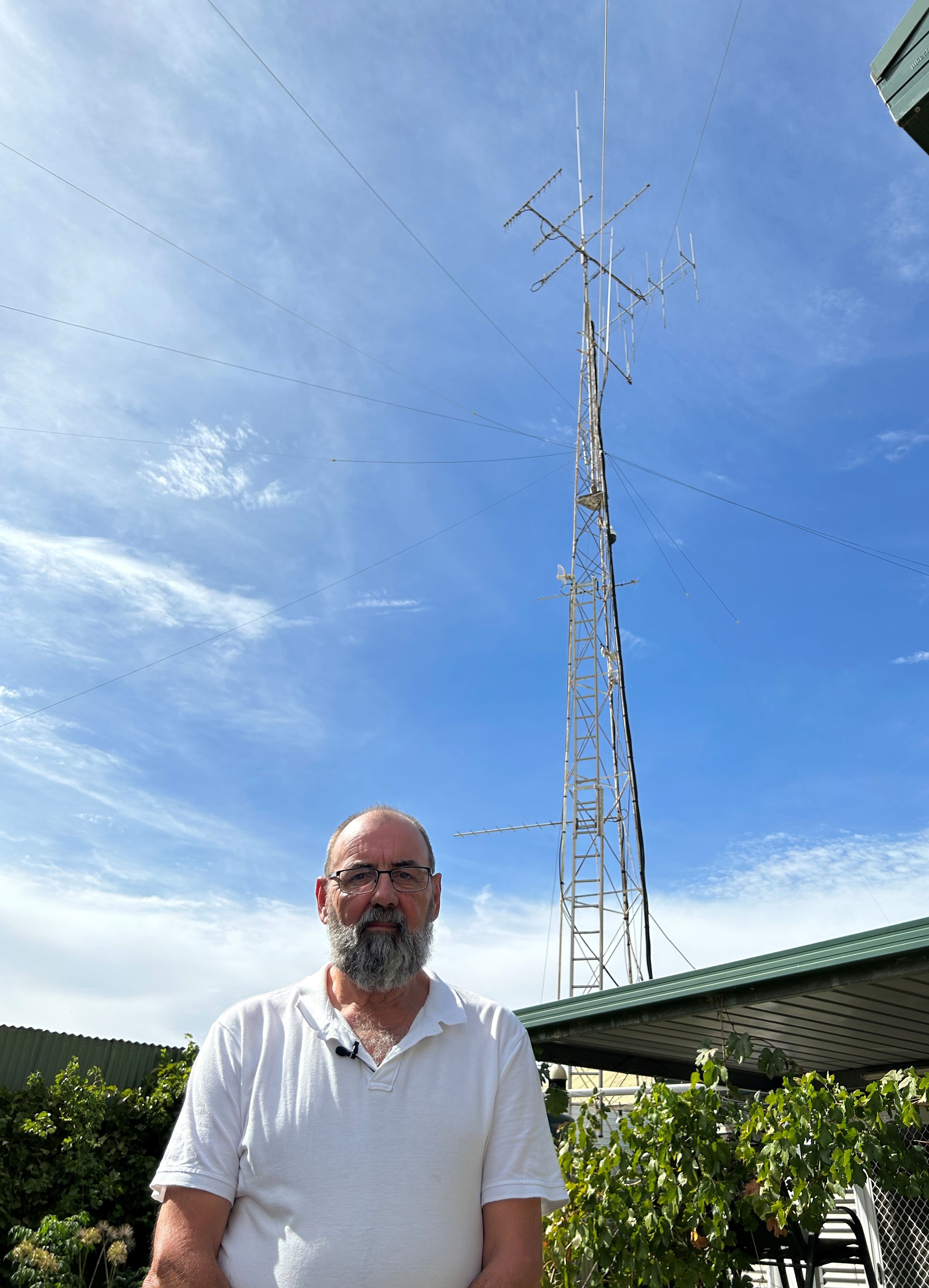A bearded man outdoors in a white shirt beneath a slender TV tower.