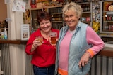 Two women laughing and standing at the bar of a pub holding a beer each. 